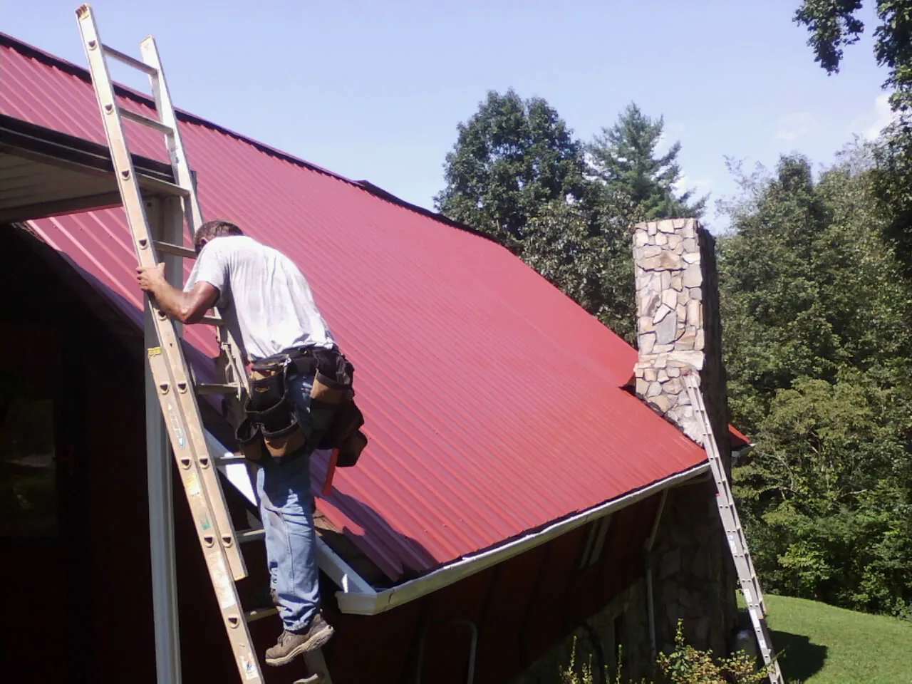 Roofer on a ladder beside a red metal roof and stone chimney — Soffit & Fascia Repair by Roofing Brothers in Forest Falls
